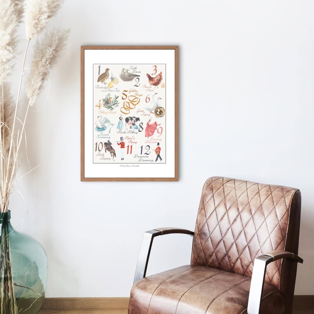 12 Days of Christmas watercolor art print framed and displayed on a wall by a leather chair and a large vase of dried flowers.