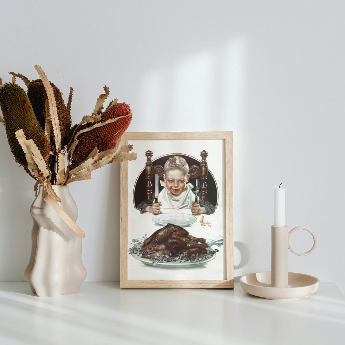 Vintage illustration of a boy excited to eat his turkey dinner, framed and displayed next to a vase with dried flowers and a candle stick.