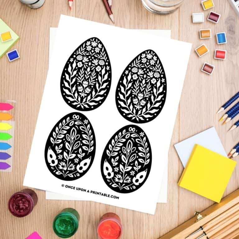 Black and white folk art Easter egg coloring pages laying on a table with coloring supplies.