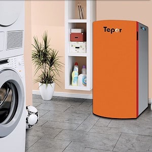 Een CV Pelletketel Tepor Compact 14-32kW in de kleuren oranje en wit staat in een wasruimte met een witte wasmachine, droger, installatie, voetbal, planken met schoonmaakmiddelen en een grijze tegelvloer.