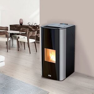 Een CV Moederhaard Tepor Steel hydro DD 14-32kW pelletkachel met glazen deur verbetert een minimalistische woonkamer. Zichtbare vlammen binnenin creëren sfeer. In de buurt staan een houten eettafel en stoelen tegen een neutrale muur, met een lichtgekleurd tapijt dat de houten vloer gedeeltelijk bedekt.