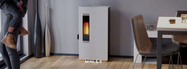 Een moderne woonkamer toont een witte Mini 5kw pelletkachel tegen een grijze muur. Een man tilt een kind op bij een groot raam. Minimalistisch decor siert de ruimte, met onder andere een donkere eettafel en bruine stoelen. "MINI 5.S" staat in het midden onderaan.