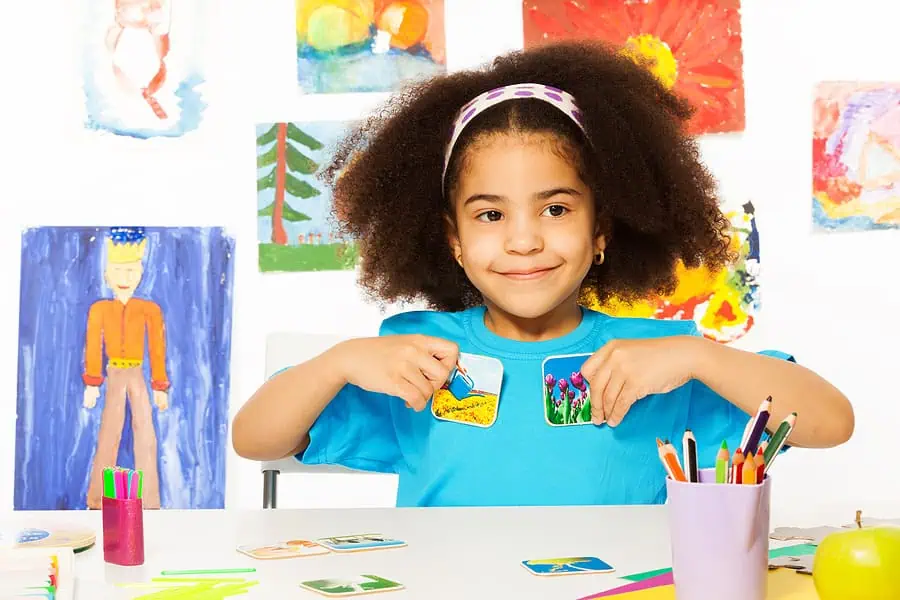 Young girl with curly hair holds flashcards at a table, surrounded by art supplies and colorful drawings on the wall.