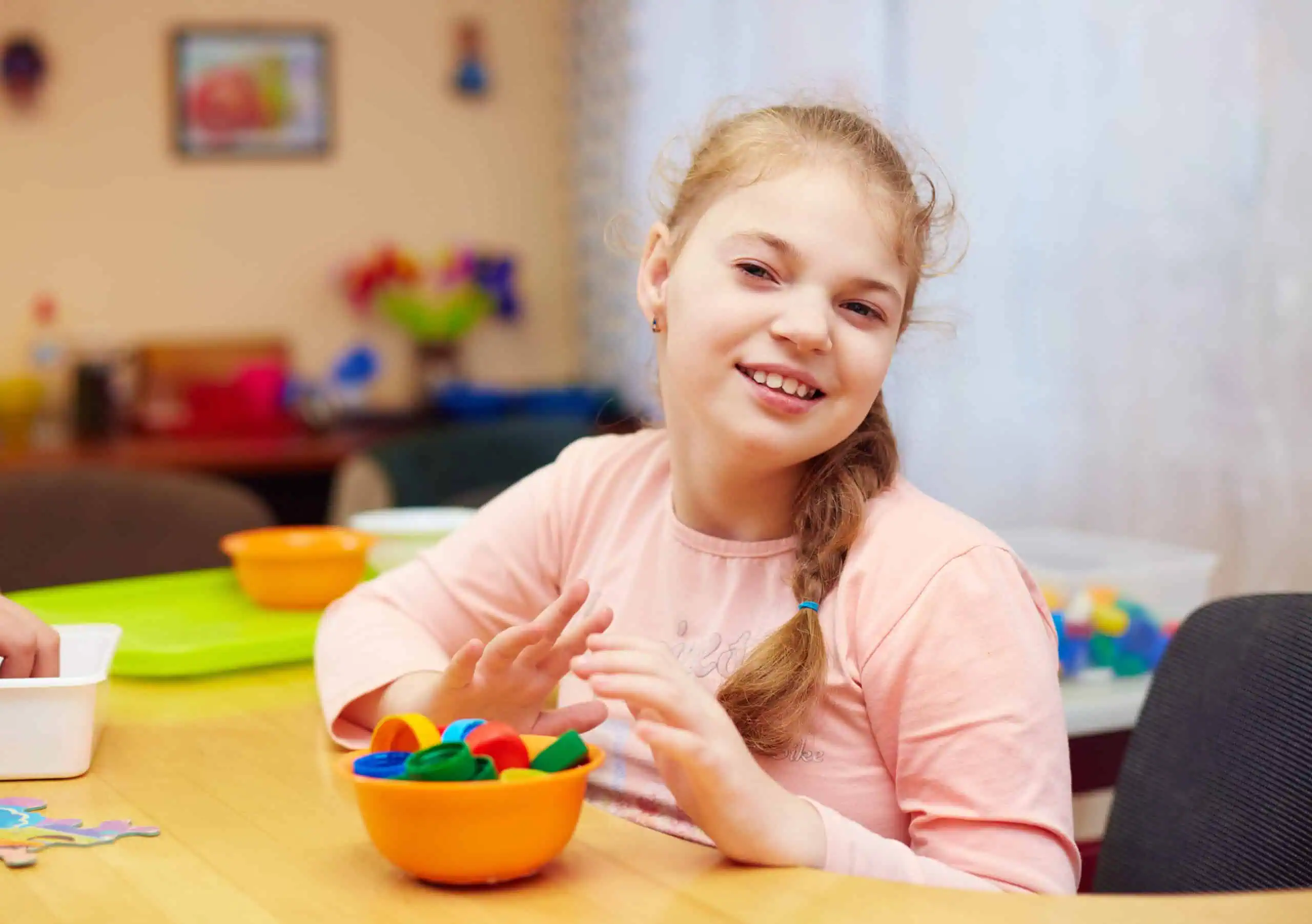 Young girl with curly hair holds flashcards at a table, surrounded by art supplies and colorful drawings on the wall.