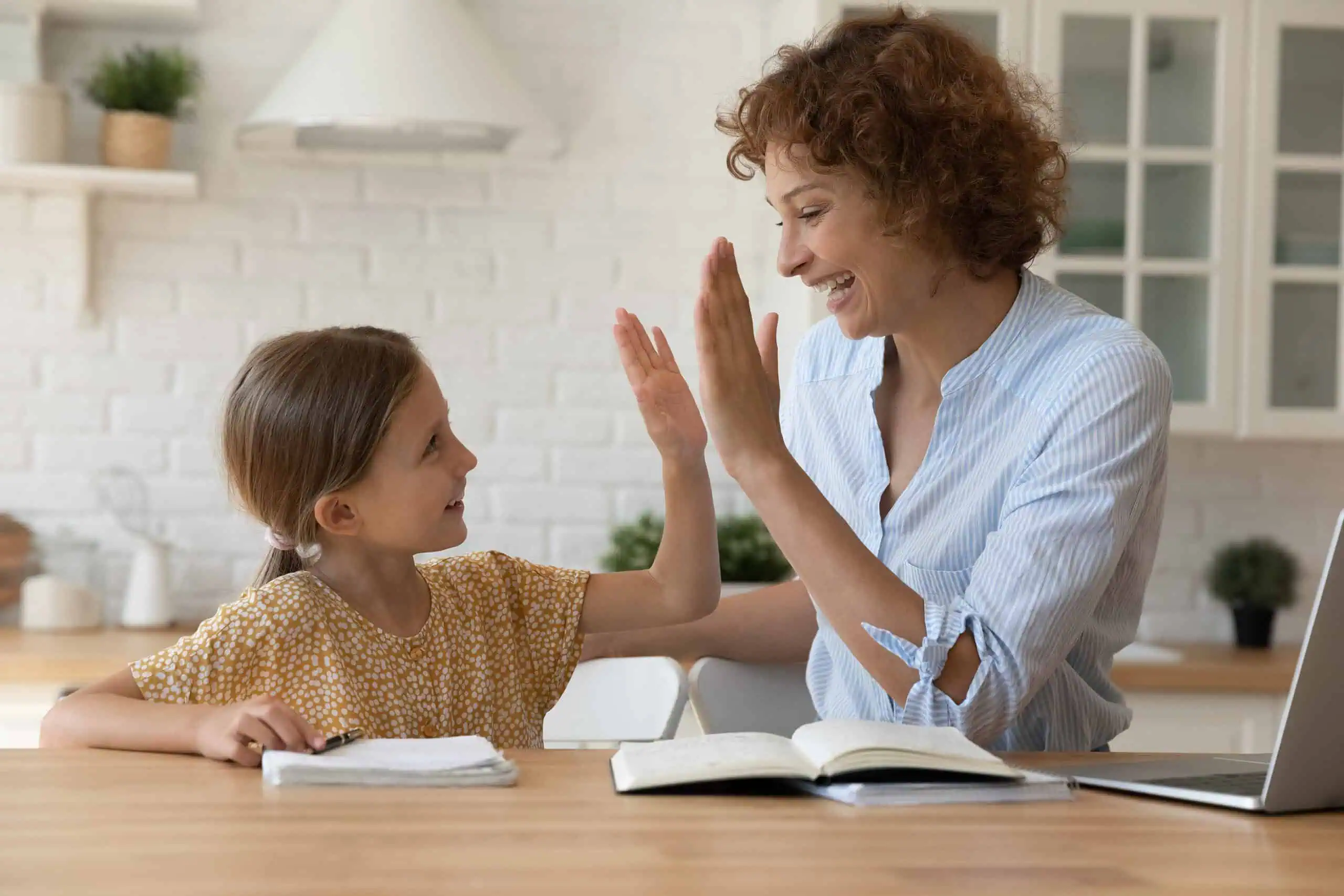 A woman and a young girl sit at a kitchen table high-fiving, with open notebooks in front of them.