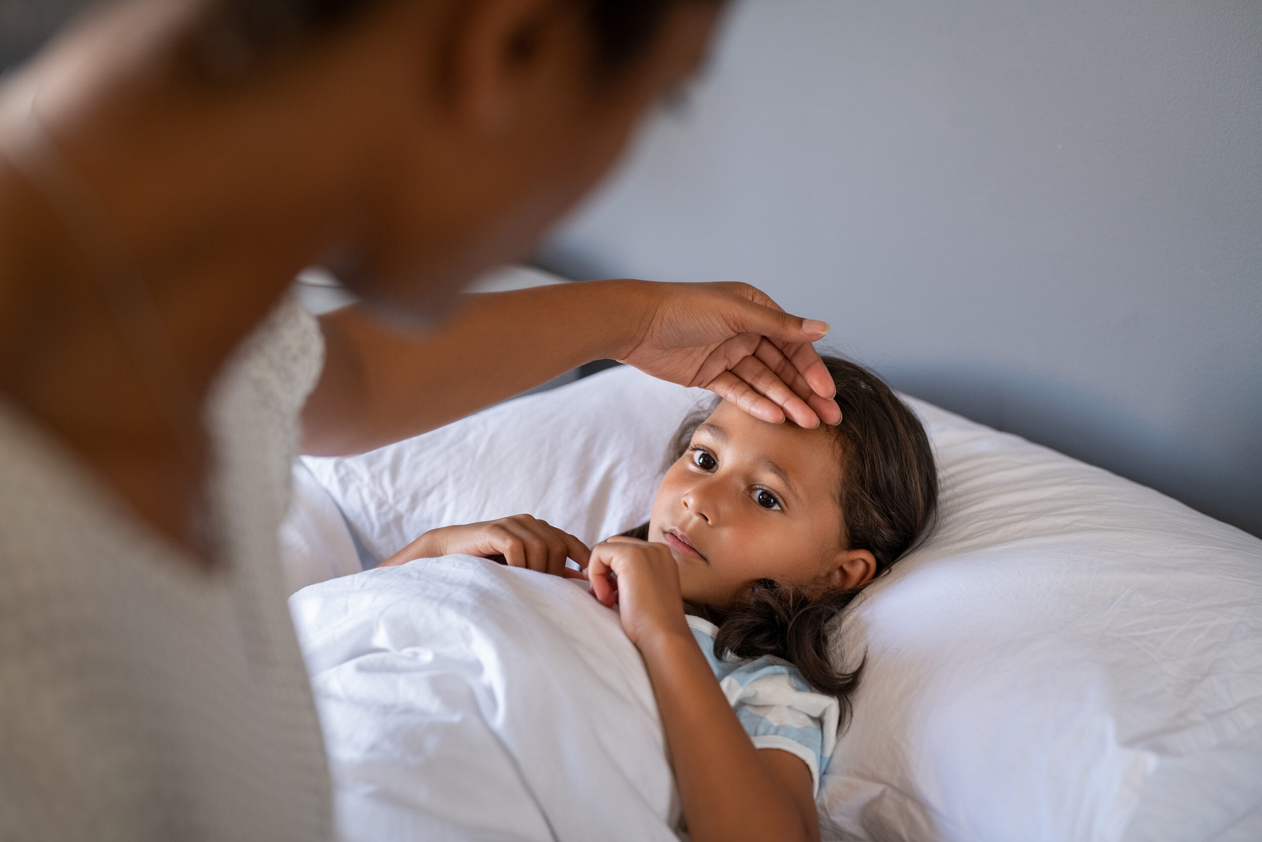 An adult checks the forehead of a child lying in bed, possibly measuring for fever. The child is covered with a white blanket, looking upward.