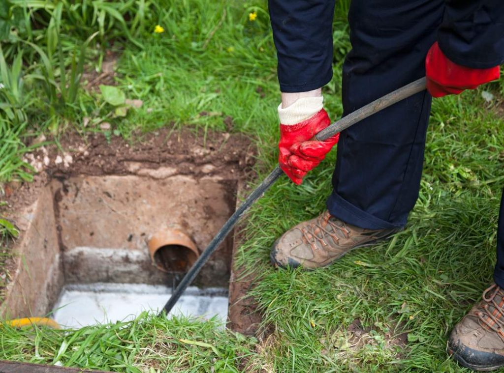 A person wearing red gloves and work boots uses a rod to clear a blocked outdoor drain, highlighting the importance of regular cleaning and maintenance for effective sewer and drain repair.