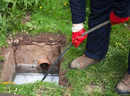 A person wearing red gloves and brown boots is using a tool to clean or inspect a pipe, showcasing professional sewer and drain repair in an outdoor drain surrounded by grass.