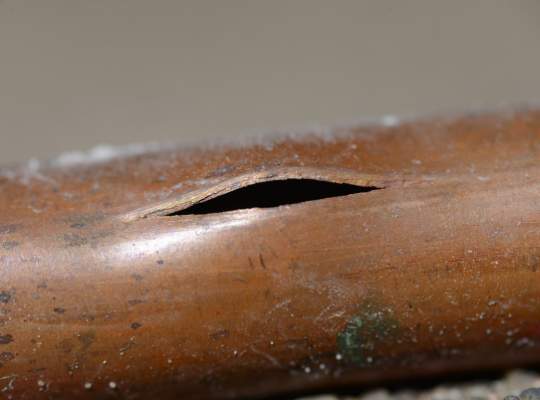 A close-up of a copper pipe with a large split or crack in its surface, exposing a dark opening inside the pipe—highlighting the need for professional pipe services or cleaning and maintenance.
