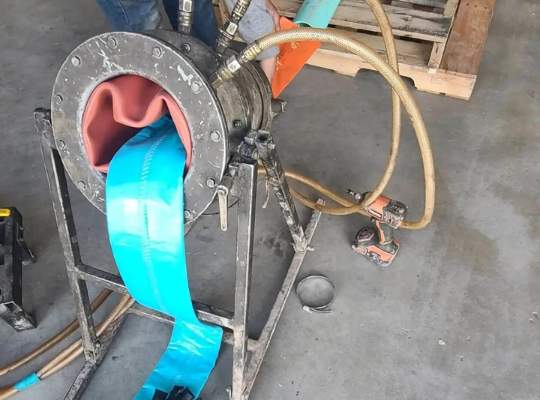 A close-up of industrial machinery extruding a blue liner for trenchless sewer repair, with hoses attached and a worker’s legs visible in the background on a concrete floor. Nearby are tools and a wooden pallet, ready for efficient pipe services.