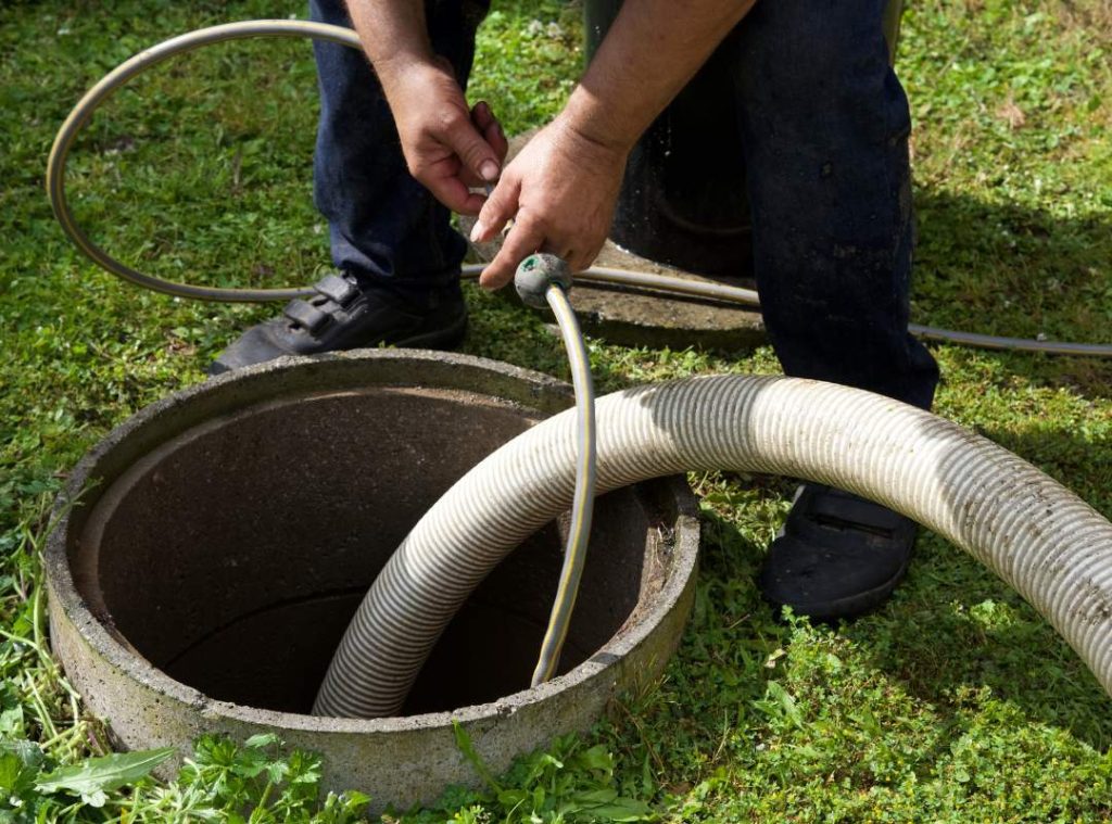 A person stands next to an open septic tank outdoors, handling hoses used for pumping and cleaning. The grassy ground around the tank highlights essential pipe services or sewer and drain repair work underway.