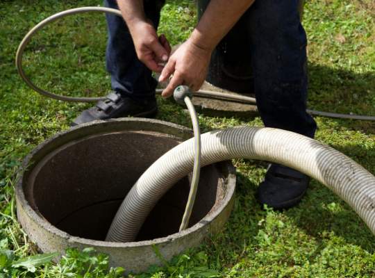 A person in dark clothing holds a hose and tube over an open outdoor septic tank, preparing for cleaning and maintenance. The scene is set on a grassy area, highlighting essential sewer and drain repair tasks.