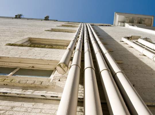Several large, white pipes run vertically up the side of a light brick building, possibly used for trenchless sewer repair or cleaning and maintenance, viewed from ground level looking upwards toward a clear blue sky.