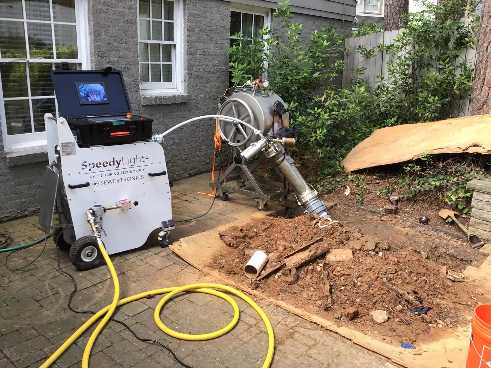 A trenchless sewer repair machine labeled “SpeedyLight+” is set up outside a house, with hoses connected to a large metal device aimed at exposed soil and pipes—showcasing advanced sewer and drain repair pipe services.