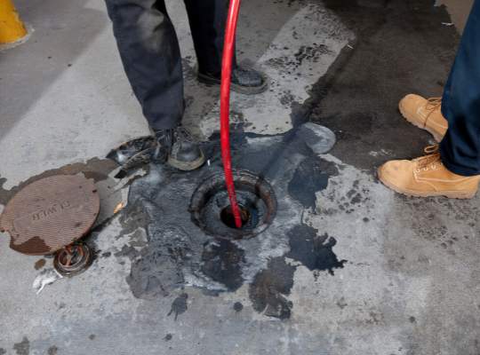 Two people stand near an open manhole cover on a concrete surface, with a red hose inserted into the hole—part of a trenchless sewer repair or cleaning and maintenance process. The area around the manhole is stained with dark liquid.