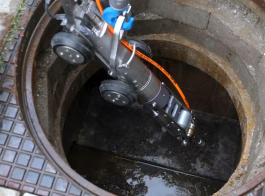 A robotic device with wheels and cables is being lowered into a circular manhole, likely for cleaning and maintenance or inspection of the underground sewer and drain repair system.