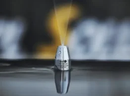 A close-up of a metal nozzle spraying a thin jet of water upward while partially submerged in a reflective surface, illustrating advanced trenchless sewer repair techniques with a blurred background.