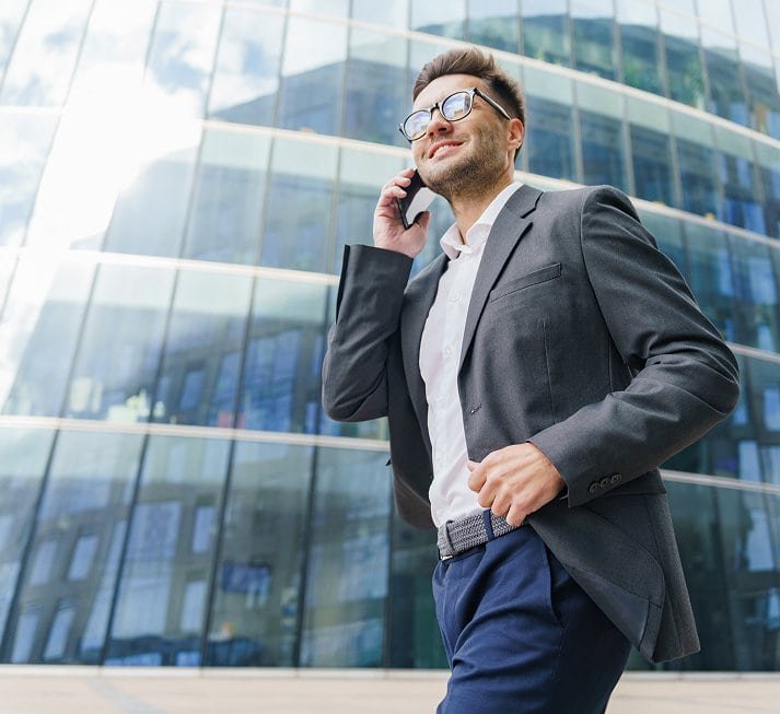Hombre con traje caminando con confianza mientras habla por teléfono frente a un moderno edificio de oficinas. Representa el acceso al mercado laboral, la comunicación profesional y el crecimiento en la carrera.
