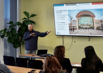 Michael Burrows, CEO, of SBD International Airport pointing to a TV screen with an airport powerpoint speaking to attendees at the Arrowhead Business Summit