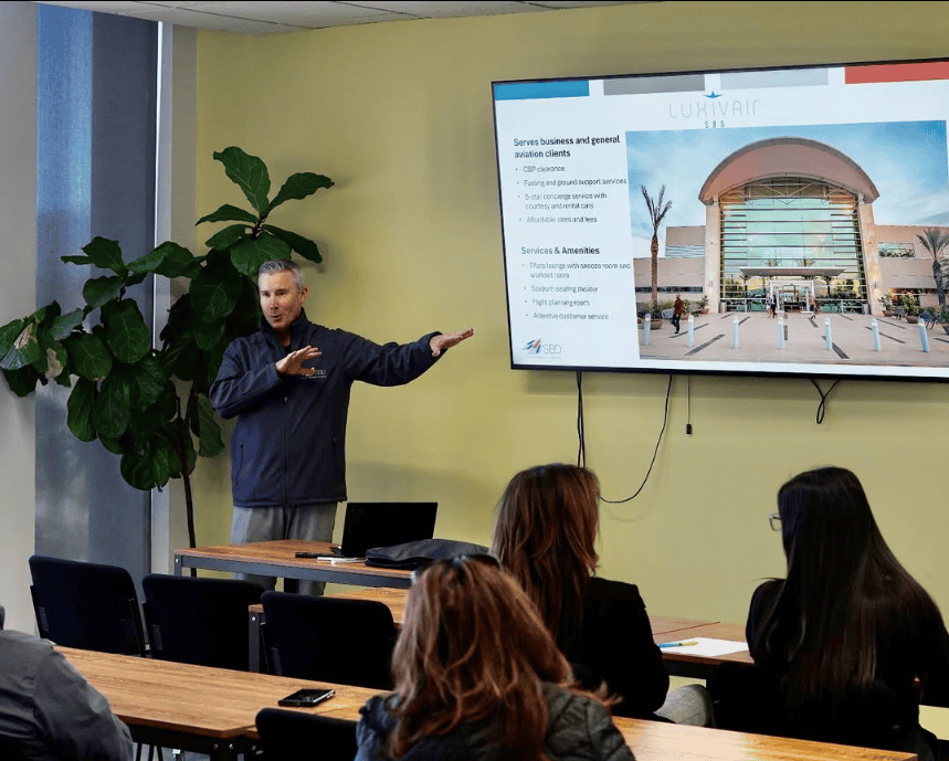 Michael Burrows, CEO, of SBD International Airport pointing to a TV screen with an airport powerpoint speaking to attendees at the Arrowhead Business Summit