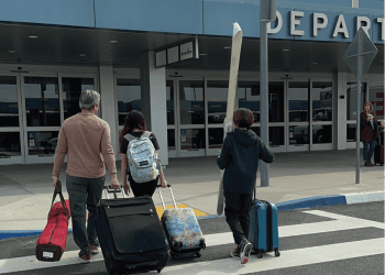 A father, son and daughter walking to the San Bernardino International Airport terminal with luggage and snow skis. A woman in standing at the curb with luggage and a cup of coffee.