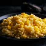 Close-up photo of fluffy, light golden-brown rice on a rustic black plate. Vibrant yellow corn kernels are mixed throughout the rice. A small pat of butter melts on top. Natural, warm lighting, emphasizing the texture and colors.