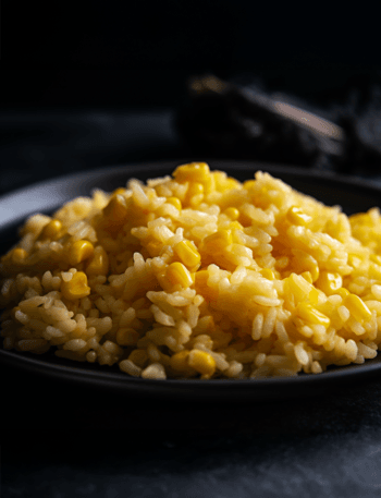 Close-up photo of fluffy, light golden-brown rice on a rustic black plate. Vibrant yellow corn kernels are mixed throughout the rice. A small pat of butter melts on top. Natural, warm lighting, emphasizing the texture and colors.