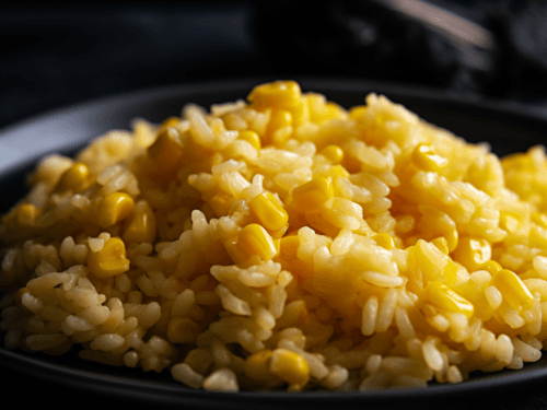 Close-up photo of fluffy, light golden-brown rice on a rustic black plate. Vibrant yellow corn kernels are mixed throughout the rice. A small pat of butter melts on top. Natural, warm lighting, emphasizing the texture and colors.