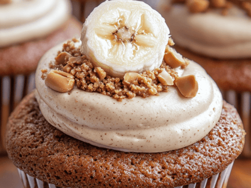 A mouthwatering photo of two perfectly baked Gluten-Free Vegan Peanut Butter Banana Cupcakes topped with swirls of fluffy Peanut Butter Cocowhip Frosting. The cupcakes are golden brown and slightly glistening, sitting on a rustic wooden board with scattered banana slices and chopped peanuts. Natural light illuminates the scene, highlighting the texture and color of the cupcakes.