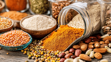 Close-up shot of various gluten-free and vegan pantry ingredients: colorful spices in small bowls, a jar of almond flour, lentils in a glass container, nuts and seeds spilling onto a wooden table