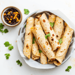 flat lay food photo of assembled taquitos on a parchment-lined air fryer basket, ready to cook. Include a small bowl of black bean filling and a pile of corn tortillas