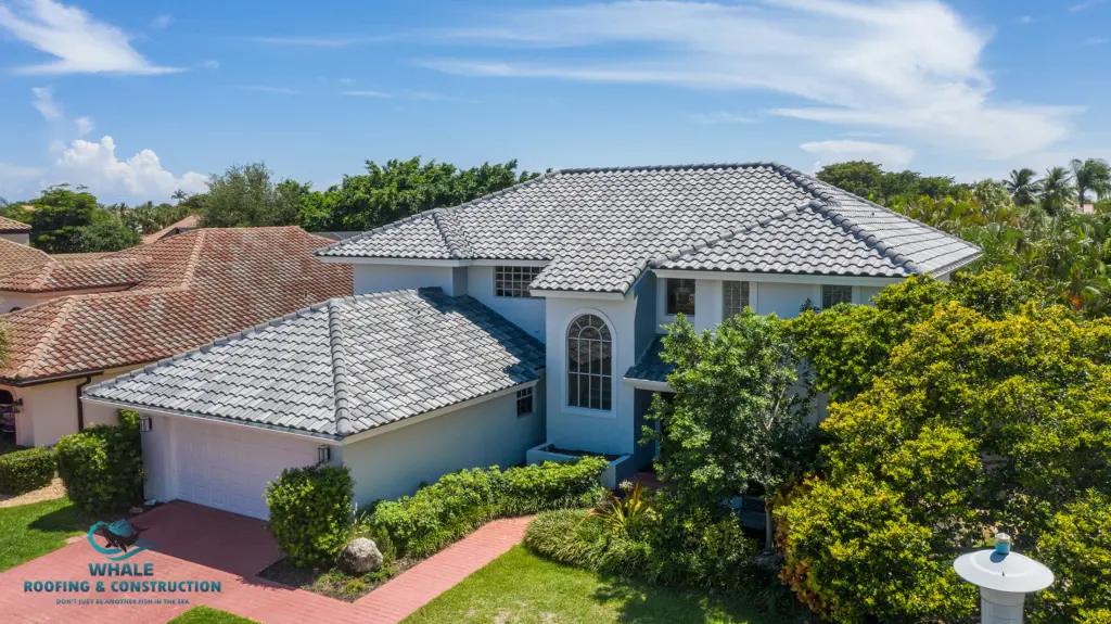 A two-story light blue house with grey tiled roof, surrounded by trees and shrubs, with a red brick driveway and a Whale Roofing & Construction logo in the corner.