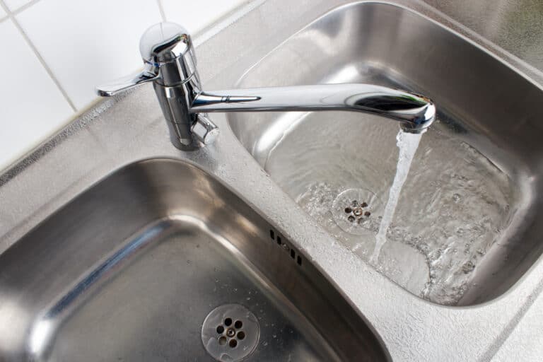Water flows from a silver faucet into a double stainless steel kitchen sink, filling the right basin while the left remains empty, ready for garbage disposal installation.