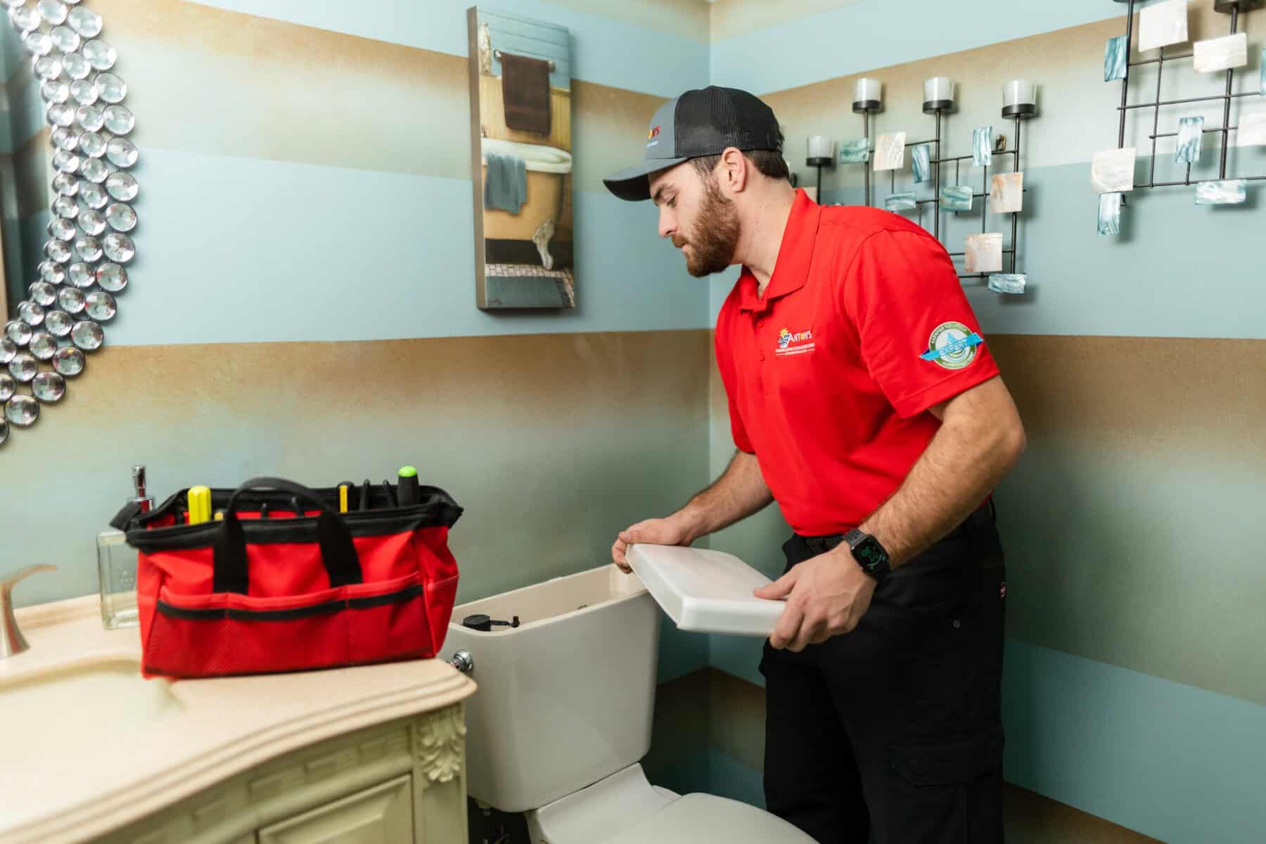 A plumber in a red shirt and cap is expertly fixing a toilet tank, tackling common plumbing problems efficiently. A tool bag rests on the sink, ready to help save money by preventing larger issues.