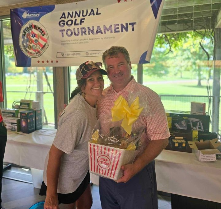 Two people smile and pose in front of a banner for an annual golf tournament. Anton, holding a gift basket labeled "Popcorn," celebrates his win as part of the Hero Program.