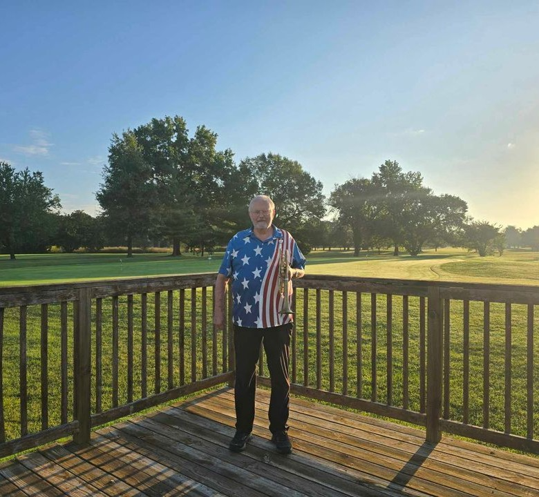 An older man named Anton stands on a wooden deck, wearing an American flag-themed shirt. Behind him, a grassy field and trees stretch out under a clear sky—a true participant in the Hero Program spirit.
