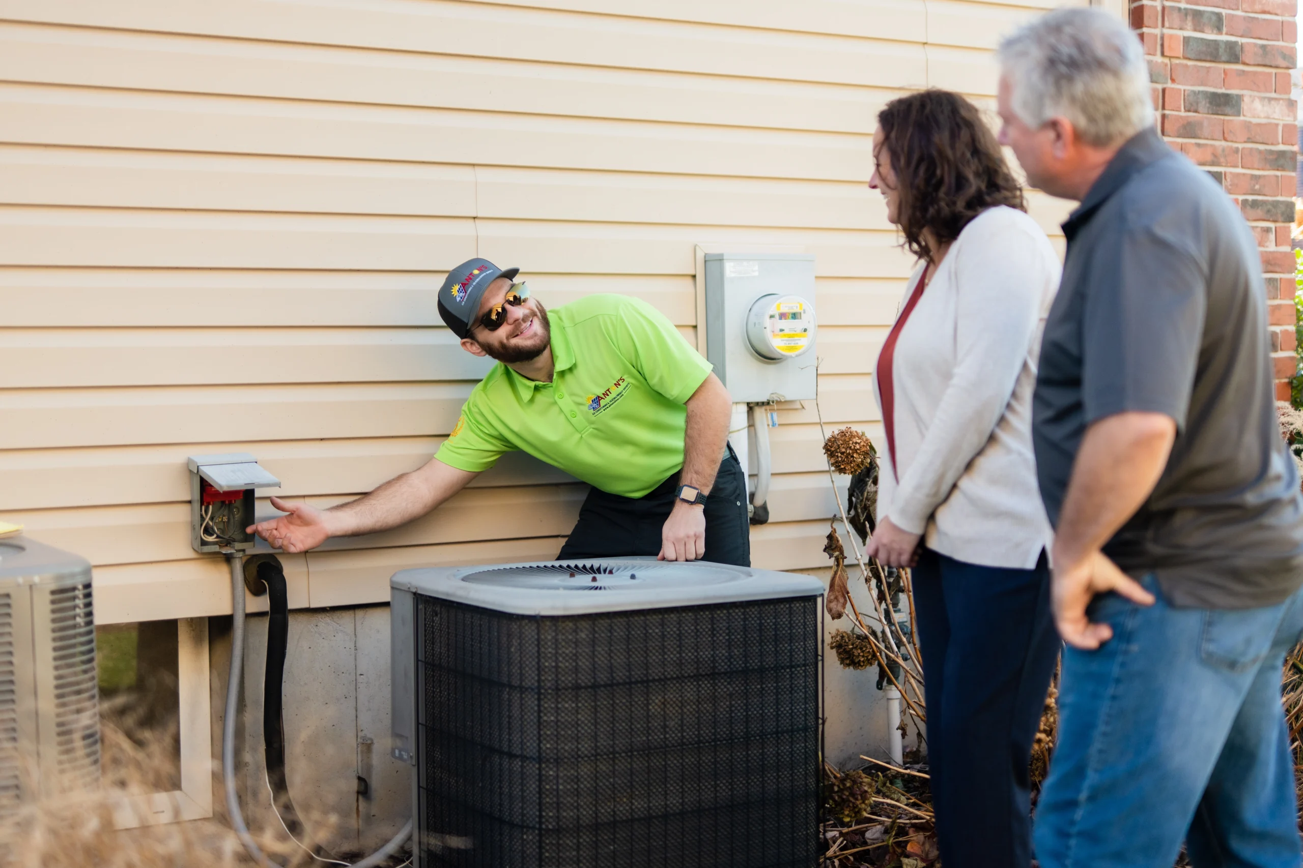 An Anton's HVAC technician explains an outdoor AC unit to a man and woman standing next to a house, discussing the importance of an air conditioner tune-up and maintenance for summer comfort in St Louis, Missouri.