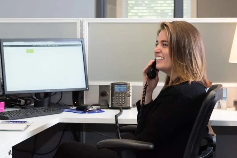 A person sits at a desk in an office, smiling while talking on a contact call via a landline phone, with a computer monitor and office equipment nearby.