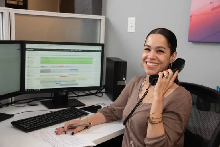 A person sits at a desk, smiling as they hold a phone in one hand. They're engrossed in work, using dual monitors to display spreadsheets while keeping a pen poised over a notepad, ready to jot down important contacts.