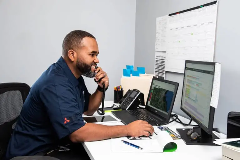 A man in a blue shirt sits at a desk, using a landline phone to contact colleagues while his eyes dart across dual monitors filled with spreadsheets. Office supplies and documents are scattered on the desk, creating an organized chaos that is the hallmark of productivity.