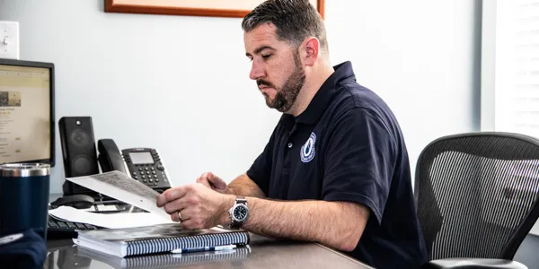 Man seated at a desk, examining a document. The cool breeze from the air conditioning installation provides comfort as he works. Computer monitor, phone, and office supplies are visible.