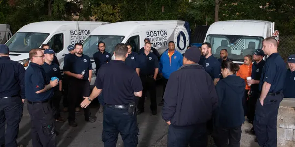 A group of people in uniforms stands in a circle outdoors near several branded vans with "Boston Standard Company" visible on them, ready for their air conditioning maintenance work. Trees provide a serene backdrop to their gathering.
