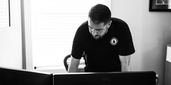 In a dimly lit office, a man leans over a desk, intently focused on two computer screens. His workspace is surrounded by tools and blueprints from a recent electrical installation, adding complexity to his concentrated endeavor.