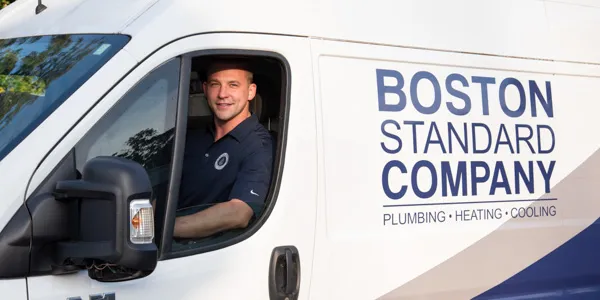 A man sits in the driver's seat of a Boston Standard Company van, prominently displaying signage for plumbing, heating, cooling, and home generator installation services.