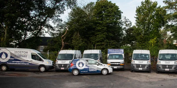 A fleet of white service vans and one car, all branded with "Boston Standard Company," is parked in the lot surrounded by trees, ready to provide premium outlets service across the city.