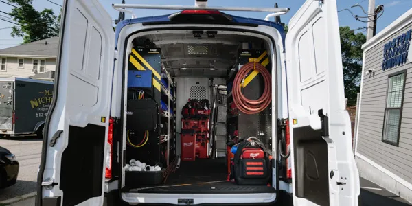 Open back of a utility van, meticulously organized for boiler maintenance with tools and equipment, including hoses and storage bins, parked on a street with buildings in the background.