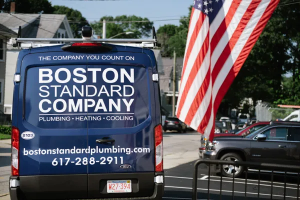 The Blue Boston Standard Company van, adorned with the company name, contact details, and services like boiler repair, is parked with an American flag waving proudly in the background.