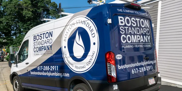 Blue and white van with Boston Standard Company branding, offering expert plumbing, heating, cooling, and furnace maintenance services. Contact information and logo with a distinct flame icon prominently displayed on the side.