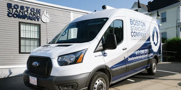 A branded Boston Standard Company van, ready for furnace repair, is parked outside a building adorned with the company's signage on the wall.