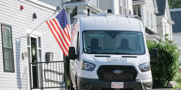 A white van with "Boston Company" on its side is parked in a residential area beside a building with an American flag, ready to provide expert heating maintenance services.